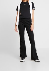 Svart T-shirt med logotyp, tillsammans med svarta utsvängda byxor med vita sidremsor. Ovanpå det bärs svarta och vita sneakers.