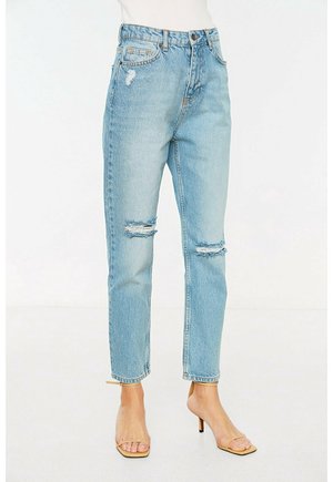 Lichtblauwe jeans met hoge taille en scheuren op beide knieën, gedragen met beige open teensandalen met hoge hak, tegen een effen witte achtergrond.