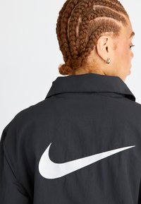 Juodas džemperis su apykakle ir dideliu baltu "Nike" swoosh logotipu ant nugaros. Audinys atrodo lygus, su minimaliu tekstūros detalėmis.