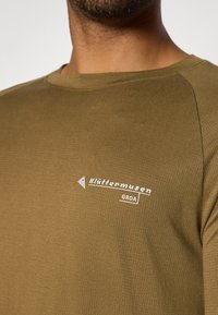 Olivengrønt skjorte med et struktureret stof, der har et hvidt "klättermusen" logo og "GROA" tekst på venstre side. Rund halsudskæring, afslappet pasform.