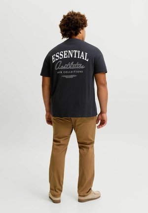 Muž s kučeravými vlasmi v čiernom tričku s nápisom „Essential Aesthetics Our Collections“ na chrbte, hnedé nohavice a béžové topánky, stojaci.