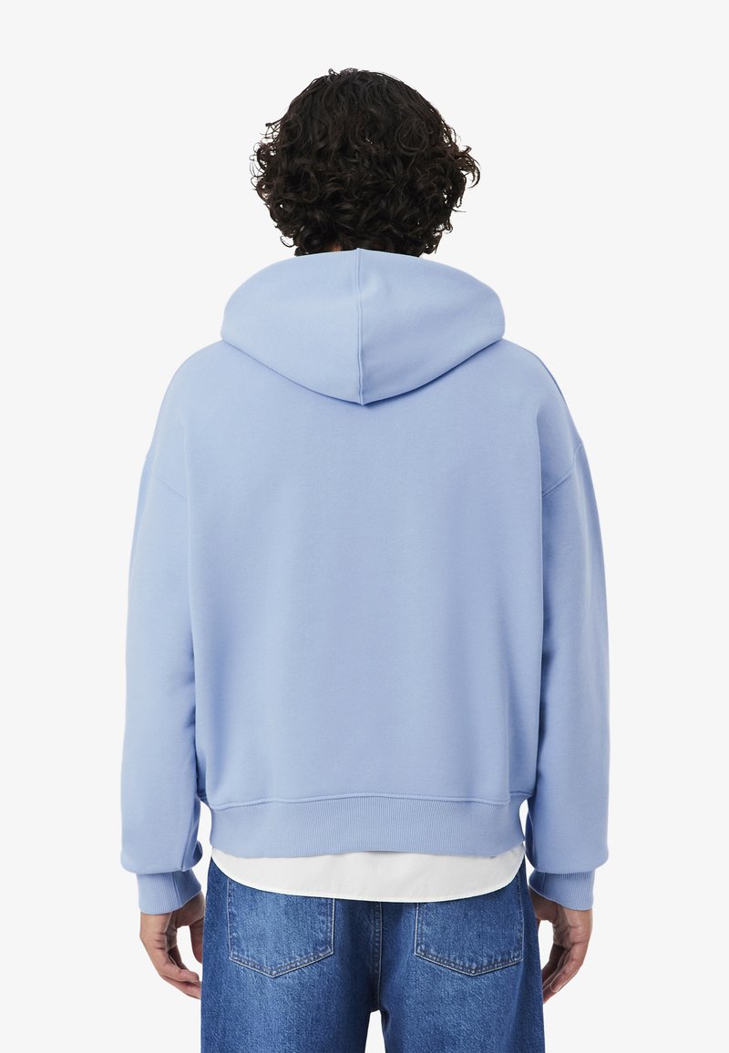 Persona con capelli ricci che indossa una felpa con cappuccio azzurro chiaro sopra una maglietta bianca e jeans blu, vista da dietro su uno sfondo semplice.