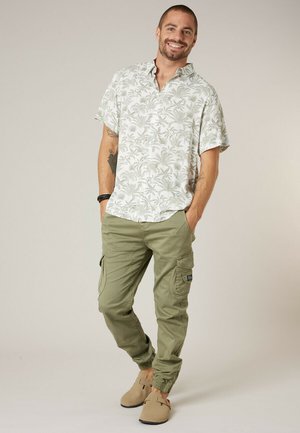 Lachende man staand met handen in zakken, gekleed in een wit t-shirt met korte mouwen en tropische print, olijfgroene cargobroek en beige instapschoenen.