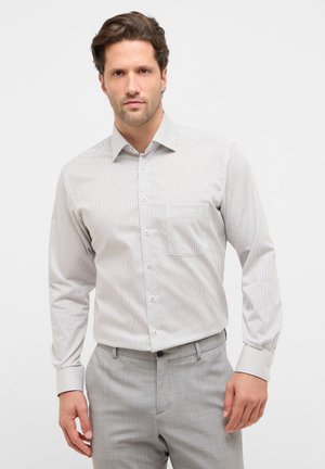 Camicia bianca a maniche lunghe con sottili righe verticali blu, colletto con bottoni, taschino sul petto e tessuto strutturato, abbinata a pantaloni grigio chiaro.