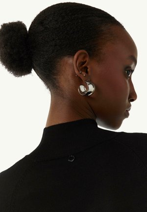 Black knit top featuring a high collar and a small circular logo at the back. Large silver earrings with a flowing design are worn.