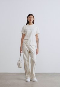 Μπλουζάκι από βαμβάκι σε κρεμ απόχρωση με λογότυπο "Calvin Klein Jeans", σε συνδυασμό με ανοιχτόχρωμο παντελόνι, λευκά αθλητικά παπούτσια και μικρή λευκή τσάντα χειρός.