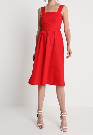 Femme portant une robe midi rouge sans manches avec un décolleté carré et des sandales blanches à talons hauts, se tenant devant un fond blanc uni.