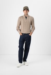 Maglione beige a maglia con mezza zip, jeans blu scuro e sneakers bianche. Modello indossa un berretto a scacchi, mani posizionate sui fianchi.