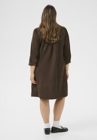 Woman with long hair wearing a loose brown knee-length dress, white socks, and black loafers standing facing away on a white background.