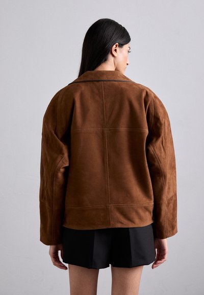 Giacca in suede marrone con un ampio colletto, spalle scese e cuciture in vista. La parte posteriore è liscia con una leggera lucentezza. Abbinata a pantaloni corti neri.