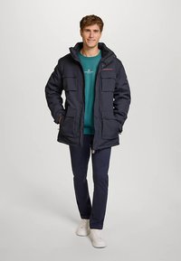 Marinblå parka med huva, flera framfickor, knapp- och dragkedjestängning, bärs över en teal tröja och mörka byxor, kombinerat med vita sneakers.