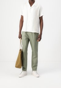 Biała koszula z krótkim rękawem i kołnierzykiem z kwiatowym haftem, zestawiona z jasnozielonymi spodniami i białymi trampkami; trzyma beżową torbę.