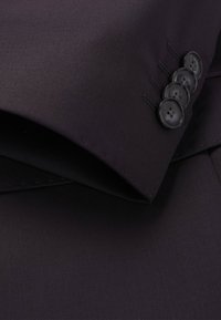 Close-up van de stof van een donkerpaarse suitjack, met een soepele textuur, nette stiksels en vier zwarte knopen aan de mouw.