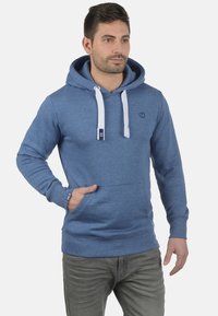 Blauwe hoodie gemaakt van zachte stof, met witte koordjes, een voorzak en een klein logo op de borst.