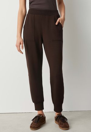Personne portant un pantalon jogger marron foncé avec des poignets élastiques et des détails de poche, associé à des baskets marron sur un fond de sol clair.