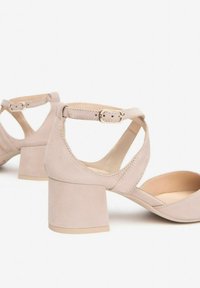 Beige suede block heel sandals with an ankle strap, open toe design, and cut-out sides. Features a gold-colored buckle detail.