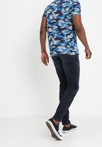 Blå kamouflage t-shirt i bomull, i kombination med mörka jeans och svarta sneakers. Fokus på bakvyn, som visar korta ärmar och en figursydd silhuett.