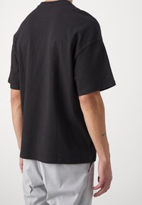 Black cotton t-shirt with short sleeves, loose fit, and crew neckline. Seen from the back, paired with light gray trousers.