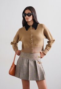 Šviesiai ruda megzta kardiganas su juodu apykakle ir perlų mygtukais, derinamas su languota mini sijonu, kuris turi auksinius akcentus ir klostes. Ruda petinė rankinė.