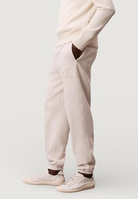 Beige joggingbroek met geborduurd logo, elastische boorden, steekzakken en een gladde textuur. Gecombineerd met lichtgekleurde sneakers.
