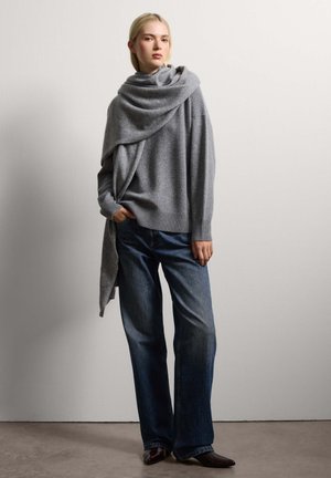 Grijze gebreide oversized trui met opgerolde halslijn en een gedrapeerd ontwerp, gecombineerd met donkerblauwe wijde jeans en puntige zwarte laarzen.