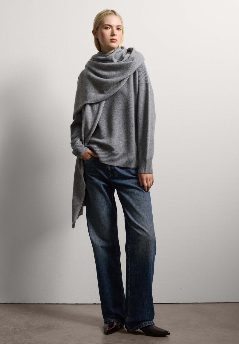 Grauer gestrickter Oversize-Pullover mit aufgerolltem Kragen und lässigem Design, kombiniert mit dunkelblauen weit geschnittenen Jeans und spitzen schwarzen Stiefeln.