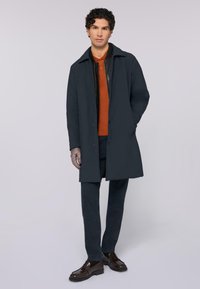 Abrigo de marino con un diseño estructurado, que incluye un cuello, botones frontales y bolsillos laterales, combinado con una polo naranja y pantalones oscuros.