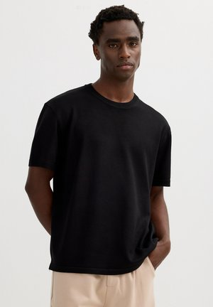 Hombre con cabello corto y rizado que lleva una camiseta negra lisa de manga corta y pantalones beige, de pie con una mano en el bolsillo sobre fondo blanco.