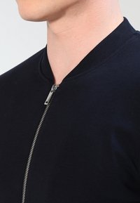 Námořnická bomberová bunda s kulatým výstřihem, stříbrným zipem a měkkou, texturovanou tkaninou. Slim-fit střih zdůrazňuje kontury.
