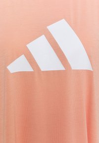 Ferskenfarvet T-shirt lavet af blødt stof med et stort hvidt Adidas-logo design med tre skrå striber på forsiden.