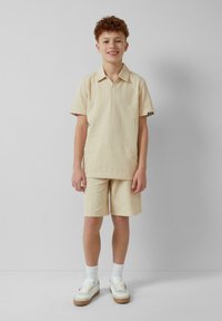 Beige gestreifte Kurzarmbluse und passende Shorts aus leichtem Material. Kombiniert mit weißen Sneakers und Socken, schlichtes Design.