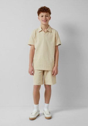 Beige gestreifte Kurzarmbluse und passende Shorts aus leichtem Material. Kombiniert mit weißen Sneakers und Socken, schlichtes Design.