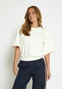 White cropped sweatshirt with a round neckline and wide sleeves, featuring a ribbed hem. Worn with navy pants that have side stripes.