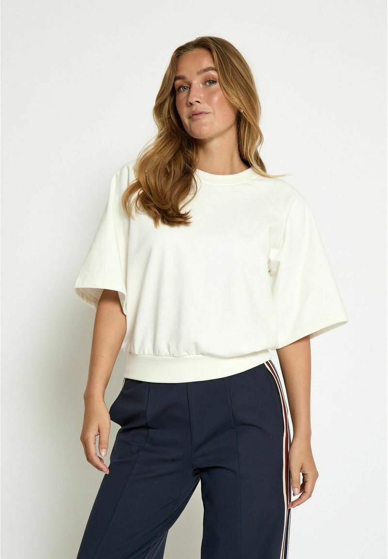 White cropped sweatshirt with a round neckline and wide sleeves, featuring a ribbed hem. Worn with navy pants that have side stripes.