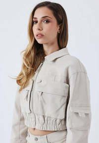 Ingekorte beige jas van lichtgewicht stof met ritsluiting, twee voorzakken, elastische tailleband en een gestructureerde kraag.