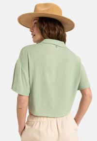 Camisa de manga corta verde claro con un acabado texturizado, cuello abotonado y corte relajado, combinada con pantalones beige.
