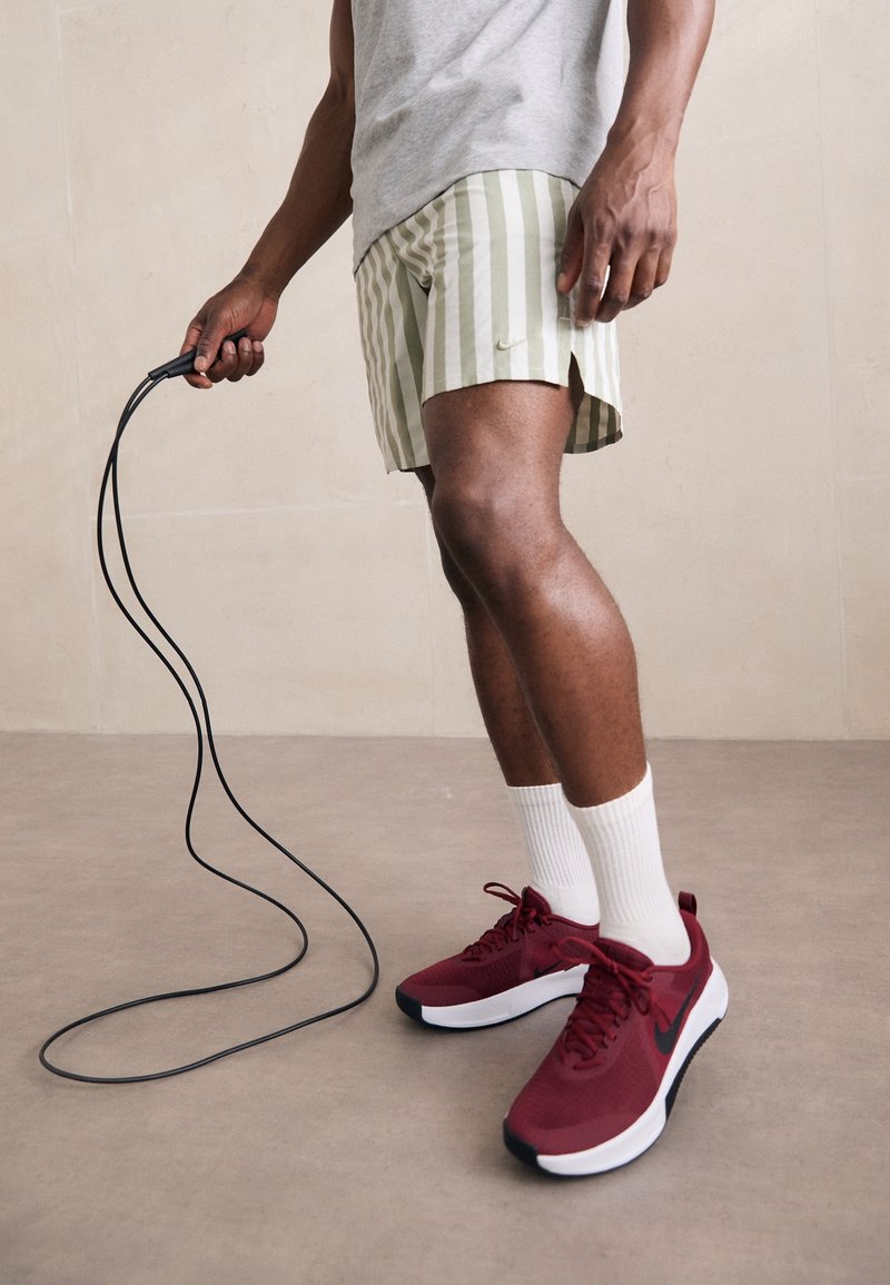 Homme portant un short rayé, un t-shirt gris, des chaussettes blanches et des baskets rouges Nike tenant une corde à sauter noire.