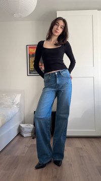 Mladá žena pózující uvnitř, oblečená v černém dlouhém rukávu, širokých modrých džínách a černých špičatých botách v prostředí ložnice.