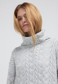 Maglione lavorato a maglia grigio chiaro con colletto alto e ripiegato. Presenta un motivo in rilievo a trecce. Design accogliente e dal taglio morbido.