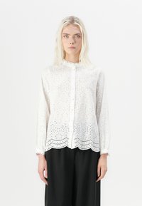 Blusa bianca a maniche lunghe con ricamo a occhio di pernice floreale, chiusura con bottoni e orlo ondulato. Tessuto morbido e leggero.