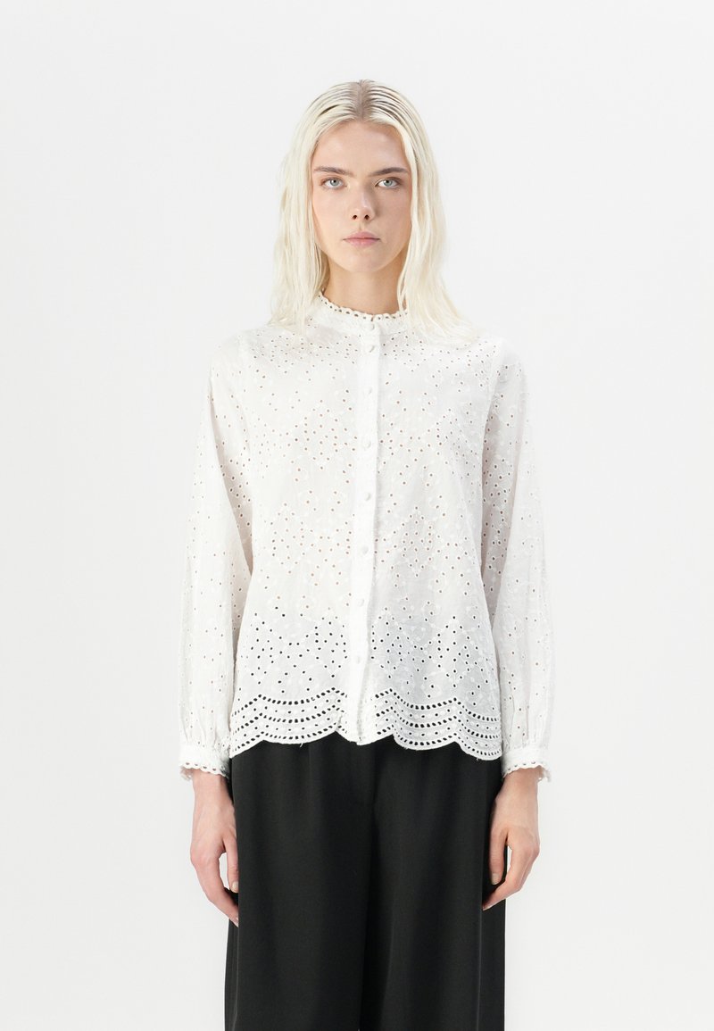 Blusa bianca a maniche lunghe con ricamo a occhio di pernice floreale, chiusura con bottoni e orlo ondulato. Tessuto morbido e leggero.