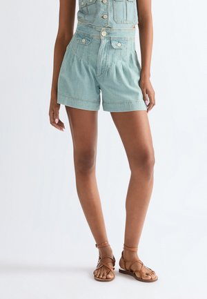 Frau trägt hellblaue High-Waist-Jeansshorts mit Taschen, kombiniert mit braunen flachen Sandalen mit Riemen, steht vor einem einfarbigen Hintergrund.