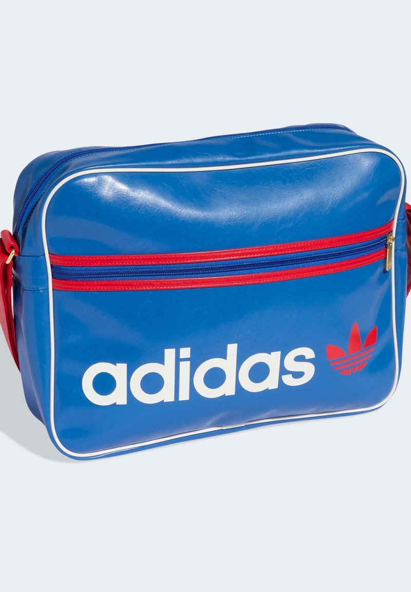 Borsa a tracolla sintetica blu con cerniera rossa, piping bianco e grande logo adidas bianco. Caratterizzata da una tasca frontale aggiuntiva con zip.