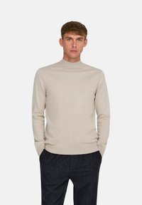 Suéter de cuello alto beige claro con una textura suave, diseño ajustado, mangas largas y puños acanalados, combinado con pantalones gris oscuro.