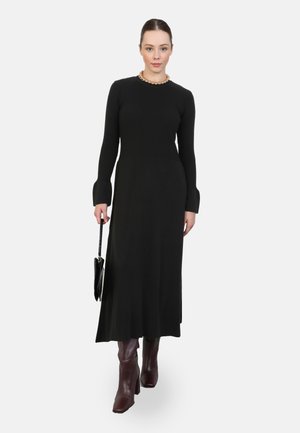 Schwarzes geripptes Strickkleid mit langen Ärmeln und ausgestellten Bündchen, kombiniert mit bordeauxfarbenen High Heels und einer schwarzen Tasche, mit rundem Ausschnitt.