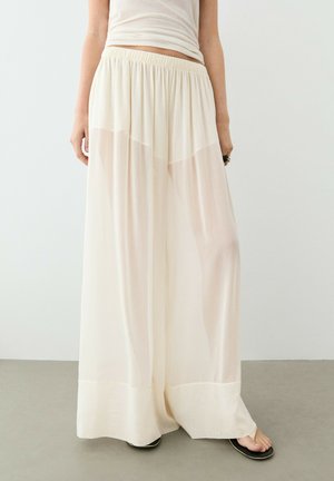 Personne portant un pantalon blanc transparent, ample à jambes larges, avec une taille élastique et des tongs noires, debout sur un sol gris contre un mur blanc.