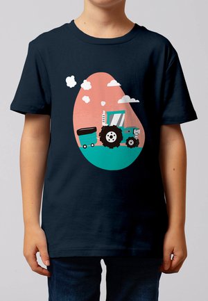 Kind trägt ein marineblaues T-Shirt mit einer Illustration eines türkisfarbenen Traktors, der einen Anhänger vor rosa-türkisfarbenem Hintergrund mit weißen Wolken zieht.