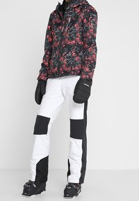 Schwarze und rote Jacke mit floralem Muster, weiße Hose mit schwarzen Flecken und schwarze Handschuhe. Outfit geeignet für Wintersport.