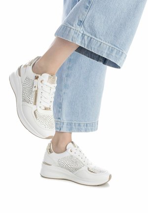 Zapatillas blancas con textura perforada, detalles en dorado y una cremallera lateral. La suela es gruesa y en forma de cuña, combinadas con unos pantalones de denim sueltos.