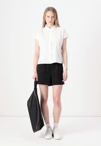 Camicia bianca a maniche corte con bottoni e motivi leggeri, abbinata a pantaloni corti neri. Il modello tiene una grande borsa a tracolla nera e indossa sneakers bianche.